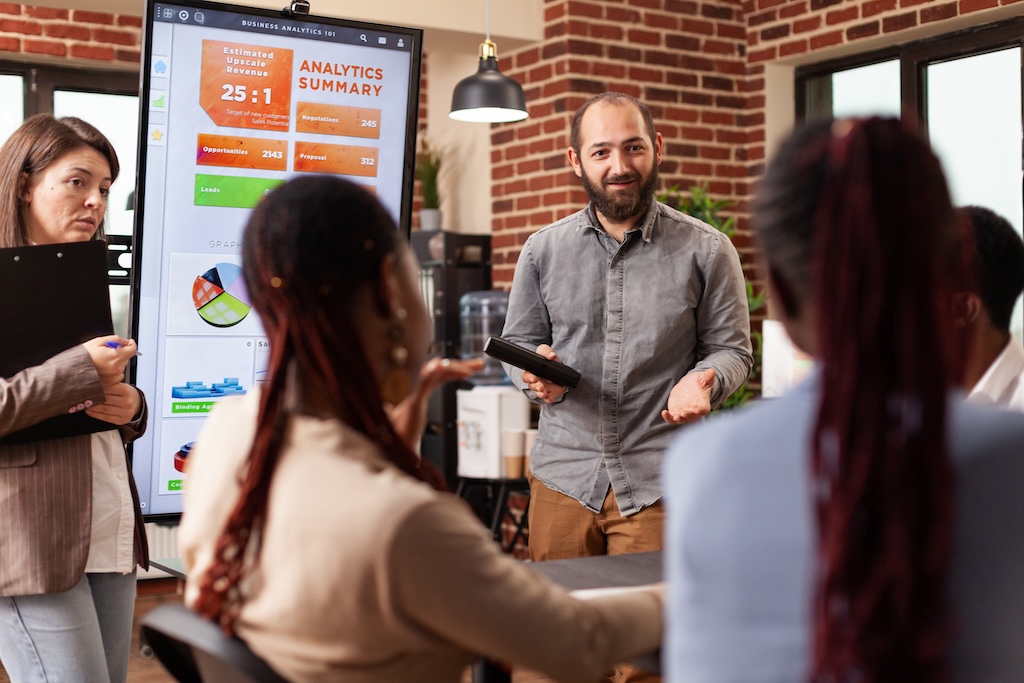 executive-manager-standing-beside-monitor-showing-marketing-graphs-explaining-company-strategy-business-meeting-startup-office-diverse-businesspeople-working-financial-presentation